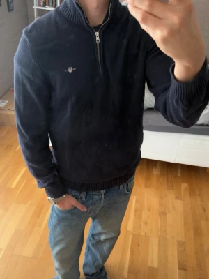 Mörkblå halvzip tröja - Mörkblå stickad tröja med halv dragkedja och ribbad krage från GANT. Tröjan har långa ärmar, ribbade muddar och en liten broderad logga på bröstet. Perfekt för lager på lager och passar till både jeans och chinos.
