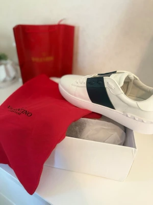 Vita Valentino sneakers med svart detalj - Snygga vita sneakers från Valentino med en bred svart läderdetalj över sidan. Skorna har klassisk låg profil, vita snören och en chunky sula med rutmönster undertill. Tillverkade i läder och levereras med originalpåse och låda.