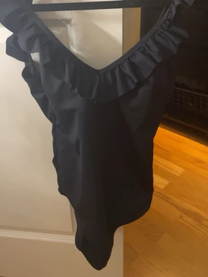 Svart baddräkt med volang, padding - Snygg svart baddräkt med djup V-ringning och volangdetaljer längs urringningen. Baddräkten har inbyggd bh för en najs look. Klassisk modell med breda axelband och stilren design. Perfekt för stranden eller poolen. Materialet är stretchigt och följsamt för en bekväm passform. Använt 1 gång.