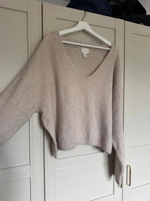 Beige stickad tröja med v-ringning - Mysig beige stickad tröja med djup v-ringning och avslappnad passform. Tröjan har långa ärmar och ribbade muddar vid ärmslut och nederkant. Perfekt för lager på lager och enkel att styla till olika outfits.