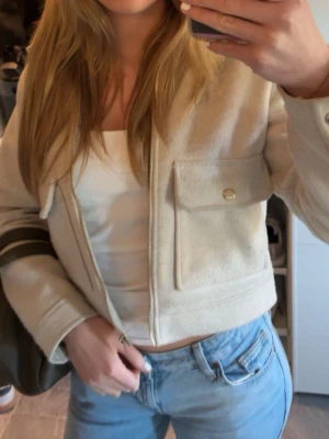 Beige cardiganjacka från ZARA - Stilren beige cardiganjacka från ZARA med krage och dragkedja framtill. Jackan har två stora fickor med lock och guldfärgade knappar. Materialet är mjukt och känns ulligt, perfekt för lager på lager under kyliga dagar.