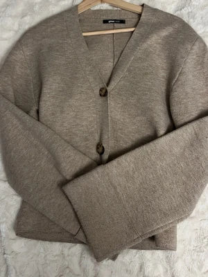 Beige kofta från Gina Tricot - Mysig beige kofta från Gina Tricot med två stora, mönstrade knappar framtill. Koftan har v-ringning, lång ärm och är stickad i ett mjukt material. Perfekt att slänga över en t-shirt eller topp för en chill look.