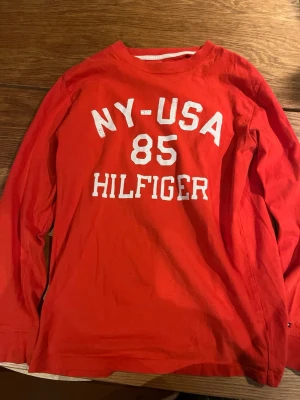 Röd långärmad topp Hilfiger - Röd långärmad T-shirt från Hilfiger med vit text 'NY-USA 85 HILFIGER' på bröstet. Klassisk rund halsringning och avslappnad passform, perfekt för en sportig och avslappnad stil.