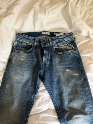 Blå slitna jeans från Replay - Säljer ett par blå jeans från Replay med snygga slitningar och tvättad look. Klassisk femficksmodell med raka ben och detaljer i kopparfärg vid fickorna. Perfekta för en avslappnad och trendig stil. Helt oanvända!