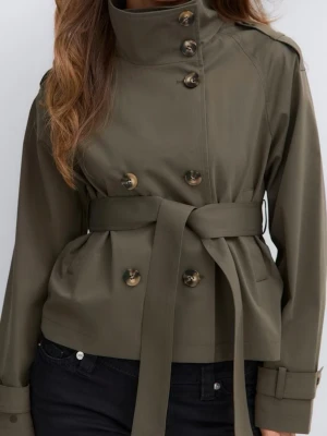 Kort olivgrön trenchcoat med bälte - Stilren kort trenchcoat i olivgrönt med dubbelknäppning och höga knappar. Jackan har ett matchande midjebälte och axelklaffar för en klassisk look. Tillverkad i ett slätt tyg med raka ärmar och justerbara ärmslut. Perfekt för dig som gillar minimalistisk och trendig stil.