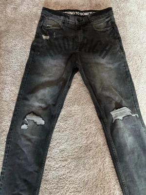 Hoodrich Jeans - Säljer ett par svarta jeans med slitningar och hål på benen. Jeansen har trycket 'HOODRICH' över framsidan och textdetalj i midjan. De har en smal passform och klassisk femficksdesign. Materialet är denim med stretch för extra komfort.