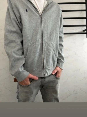 Grå hoodie från Polo Ralph Lauren - Snygg grå hoodie från Polo Ralph Lauren med dragkedja framtill och två stora fickor. Klassisk liten mörkblå logga på bröstet och mjukt material på insidan. Perfekt för en chill och avslappnad stil.