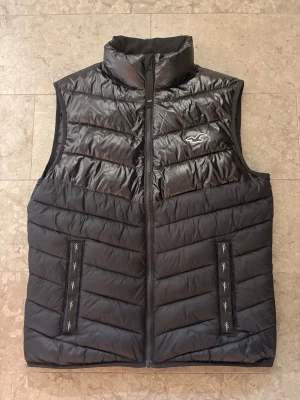 Svart dunväst från Hollister - Snygg svart dunväst från Hollister med quiltad design och hög krage. Västen har två fickor framtill med dragkedja och Hollisters ikoniska logga på bröstet. Perfekt för lager-på-lager och håller dig varm under kyliga dagar.