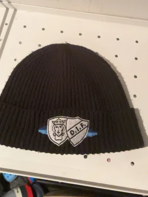 Svart ribbstickad DIF-mössa från sofialäktaren  - Svart ribbstickad mössa från sofialäktaren med broderad Djurgårdens IF-logga framtill. Mössan har uppvikt kant och detaljer i blått och vitt. Perfekt för dig som vill visa ditt stöd för laget under kalla dagar.