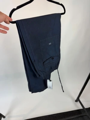 Marinblå byxor från C.P. Company - Snygga marinblå byxor från C.P. Company med elastisk midja och dragsko. Byxorna har en ficka med knapp baktill och är tillverkade i ett lätt material, perfekta för vardagsbruk eller avslappnade tillfällen. Prislapp kvar! Köp direkt eller lägg ett bud.
