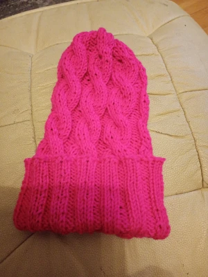 Rosa kabelstickad mössa - Säljer en snygg rosa kabelstickad mössa med uppvikt kant. Mössan är gjord i mjukt garn och har ett klassiskt flätmönster som ger en trendig look. Perfekt för att hålla värmen under kalla dagar och ger en färgklick till din outfit.