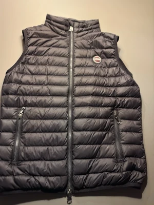 Svart barn colmar väst -  Storlek 154/ 12 år. Snygg svart dunväst från Colmar Originals med quiltad design och hög krage. Västen har två dragkedjeförsedda fickor och Colmar-logga på bröstet. Perfekt för lager-på-lager och håller dig varm under kyliga dagar. Unisex