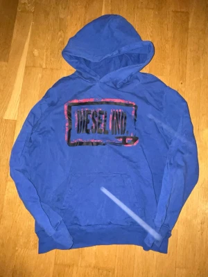Diesel hoodie - Snygg blå hoodie från Diesel med stort DIESEL IND. tryck i svart och rött på bröstet. Klassisk huva och magficka framtill. Perfekt för en avslappnad streetwear-look.