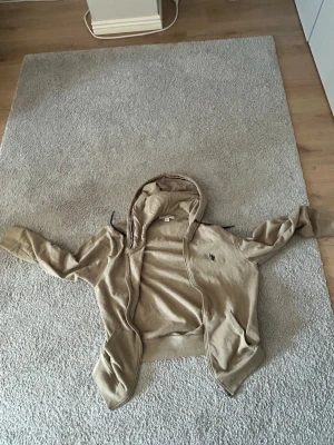 Beige hoodie med dragkedja - Säljer en beige hoodie med dragkedja och huva som jag aldrig använt. Tröjan har långa ärmar, fickor framtill och en liten broderad logga på bröstet. Perfekt för chill dagar eller när du vill ha något bekvämt och stilrent.