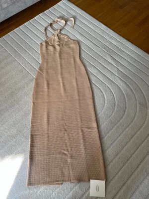Beige ribbad långklänning från Adoore - Trendig beige långklänning från Adoore med ribbad struktur. Klänningen har halterneck med knyt i nacken och en smickrande, figurnära passform. Perfekt för sommarens alla tillfällen.