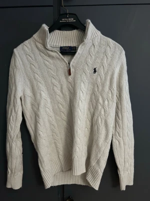Vit kabelstickad tröja Polo Ralph Lauren - Kabelstickad vit tröja från Polo Ralph Lauren med halv dragkedja och hög krage. Tröjan har ribbade muddar och den klassiska broderade loggan på bröstet. Perfekt för lager på lager och riktigt snygg till jeans. Super snygg tröja med den klassiska half zip! Pris kan diskuteras vid snabb affär 