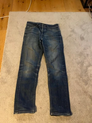 Jack and Jones jeans - Säljer ett par blå slim fit jeans med snygga slitningar och tvättade detaljer. Klassisk femficksmodell med dragkedja och knapp i midjan. Jeansen har en modern passform och är perfekta för en avslappnad stil.