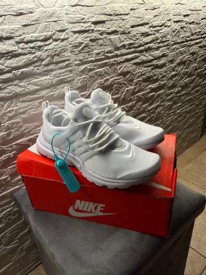 Nike vita prestos - Säljer ett par helvita Nike sneakers med mesh-överdel och plastdetaljer vid snörningen. Skorna har en sportig look med rund tå, vita snören och dragflikar bak. Sitter på en klassisk Nike-låda. Perfekta för dig som gillar stilrena och bekväma sneakers.