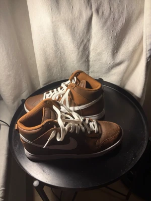 Bruna Nike sneakers med vita detaljer - Snygga bruna Nike sneakers med vita detaljer och vit sula. Skorna har en klassisk mid-modell och är tillverkade i läder med perforerad tåbox för extra ventilation. Vita skosnören och den ikoniska swoosh-loggan på sidan ger en clean look.