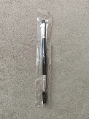Svart ögonskuggsborste med spoolie - Dubbeländad ögonskuggsborste från MAGEFY med svart handtag. Ena sidan har en vinklad borste för precis applicering och andra sidan har en spoolie för att borsta bryn eller fransar. Borsten har metallfästen och syntetiska strån.
