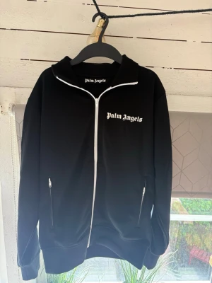 Svart hoodie från Palm Angels - Säljer en svart hoodie från Palm Angels med vit dragkedja och logga på bröstet. Hoodien har två fickor med dragkedja och en klassisk huva. Perfekt för dig som gillar streetwear och vill ha en stilren look.