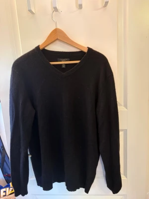 Svart kashmirtröja från Club Room - Mjuk och lyxig svart stickad tröja från Club Room i 100% kashmir. Tröjan har V-ringning och långa ärmar, perfekt för dig som vill ha en klassisk och stilren look. Passar lika bra till jeans som till chinos.