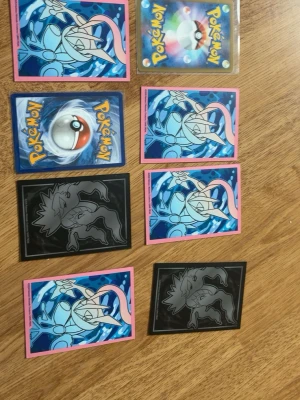 Samling Pokémonkort med sleeves - Säljer en samling Pokémonkort med olika motiv och färgglada sleeves. Inkluderar både vanliga och holografiska kort, samt skyddande plastfickor i rosa och svart. Perfekt för samlare eller Pokémonfans som vill utöka sin kortlek.