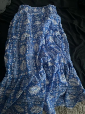 Blå långkjol med blommigt mönster - Säljer en blå långkjol med vitt blommigt mönster. Kjolen har en avslappnad passform och är gjord i ett lätt material som passar perfekt till varmare dagar. Midjan är elastisk för extra komfort.
