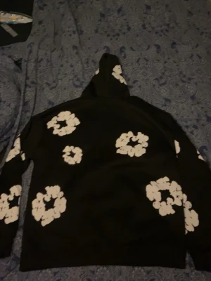 Denim tears - Säljer en svart hoodie med stora vita blommönster över hela plagget, även på huvan. Hoodien har en klassisk passform och är tillverkad i mjukt material, perfekt för en avslappnad och trendig stil.