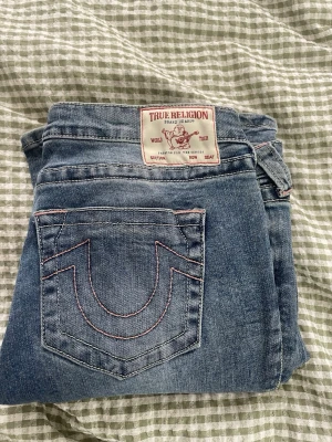 Blå True Religion jeans med kontrastsömmar - Säljer ett par klassiska blå jeans från True Religion med ikoniska kontrastsömmar i rosa och tydliga bakfickor. Jeansen har låg midja, bootcut och silverfärgade knappar. Perfekta för dig som gillar streetwear och vill sticka ut lite extra. Använda två gånger dom är tyvär för stora för mig 