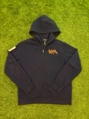 Mörkblå hoodie med polospel-tryck 18/20 år - Snygg mörkblå hoodie från Polo Ralph Lauren med dragkedja och huva. På bröstet finns ett broderat motiv med polospelare i färgglada tröjor. Hoodien har ribbade muddar och fickor framtill. Perfekt för en avslappnad och sportig stil passar parfekt om du är 175.