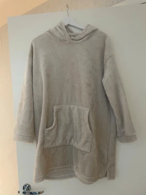 Oversized Fleece Hoodie Blanket - This is an oversized hoodie blanket made from soft, plush fleece material. It features long sleeves, a large front pocket, and a hood. The color is a light beige or cream. The garment is designed for comfort and warmth, ideal for lounging at home.
