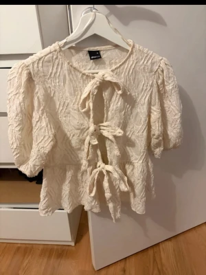 Krämvit blus med knyt framtill - Supersöt krämvit blus med tre knytband framtill och lätt puffade trekvartsärmar. Blusen har en avslappnad passform och volang nedtill, perfekt för en trendig och chill look.