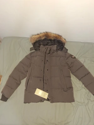 Brun dunjacka med fuskpälsluva - Snygg brun dunjacka med avtagbar luva och fuskpälsdetalj. Jackan har ribbade muddar vid ärmsluten, två sidofickor och en patch på ärmen. Perfekt för kalla dagar och riktigt varm tack vare dunfyllningen.