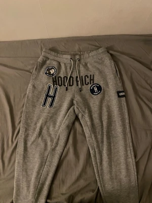 Grey Hoodrich Joggers - These are grey jogger sweatpants from the brand Hoodrich. The joggers feature multiple embroidered and printed patches, including a large 'H', 'HOODRICH' text, and circular patches with phrases like 'FROM NOTHING TO SOMETHING' and 'HOODRICH UK LIFESTYLE'. The joggers have a drawstring waistband and appear to be made from a soft, comfortable material suitable for casual wear.