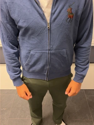 Blå zip hoodie från Polo Ralph Lauren - Snygg blå hoodie från Polo Ralph Lauren i storlek S. Hoodien har dragkedja framtill, två fickor och den klassiska broderade loggan på bröstet. Tillverkad i mjukt material som känns skönt mot huden. Perfekt för en avslappnad och stilren look.