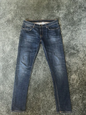 Mörkblå jeans från Nudie Jeans - Säljer ett par mörkblå jeans från Nudie Jeans med klassisk femficksmodell och orange kontrastsömmar. Jeansen är tillverkade i slitstark denim. Perfekta för dig som gillar en tidlös och clean look.