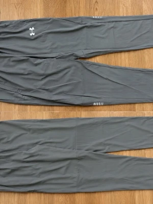 Grå träningsbyxor Under Armour - Snygga grå träningsbyxor från Under Armour med smal passform och diskreta vita detaljer. Byxorna har elastisk midja och är tillverkade i ett lätt, stretchigt material som passar perfekt för träning eller sportaktiviteter.