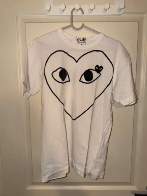 Vit COMME des GARCONS T-shirt  - Vit t-shirt från COMME des GARCONS med ett stort svart hjärta och ögon på bröstet. T-shirten har rund hals och korta ärmar. Materialet ser ut att vara bomull och modellen är klassisk och normal i passformen. Perfekt för dig som gillar grafiska tryck och unik stil. Små håll baktill, se på bild 