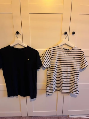 2-pack Lyle & Scott t-shirts, svart & randig - Två snygga t-shirts från Lyle & Scott i juniorstorlek. Ena är helsvart med liten gul logga på bröstet, den andra är vit med svarta ränder och samma logga. Båda har rund hals och korta ärmar. Perfekta till vardagslooken!