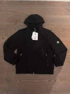 Svart vindjacka från Moncler - Snygg svart vindjacka från Moncler med huva och dragkedja framtill. Jackan har två fickor med dragkedja och Moncler-logga på ärmen. Tillverkad i lätt och slitstarkt material, perfekt för blåsiga dagar.