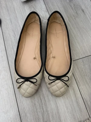 Beige quiltade ballerinaskor med rosett - Snygga ballerinaskor i beige med quiltat mönster och svarta detaljer. Skorna har en liten svart rosett framtill med metalländar och svarta kanter runt öppningen. Perfekta för en stilren och klassisk look.