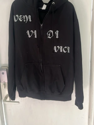 Svart hoodie med tryck från Blinglucid - Svart zip-hoodie från Blinglucid med texten 'Veni Vidi Vici' i vit gotisk stil på framsidan. Hoodien har huva med dragsko, två fickor framtill och är tillverkad i bomull och polyester. Perfekt för en streetwear-look.