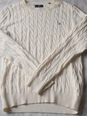 Vit kabelstickad tröja från Gant - Kabelstickad tröja i premium bomull från Gant. Tröjan är vit med rund halsringning, ribbade muddar och diskret Gant-logga broderad på bröstet. Perfekt för en clean och klassisk stil.