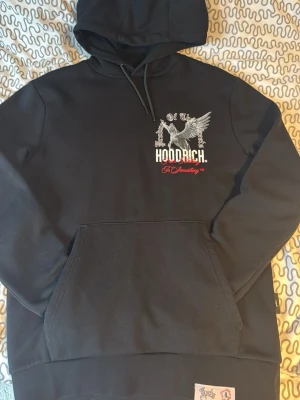 Svart hoodie från Hoodrich med tryck - Svart hoodie från Hoodrich med stor ficka framtill och coolt Pegasus-tryck på bröstet. Texten 'Product of the Streets' och 'From Nothing To Something' i vitt och rött ger streetwear-vibe. Mjuk insida och klassisk huva med dragsko.