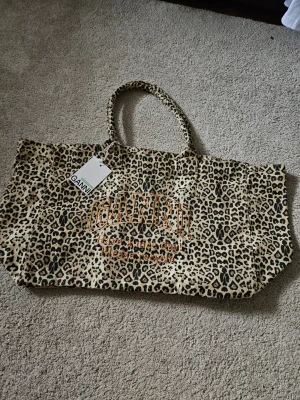 Leopardmönstrad shoppingväska från Ganni - Säljer en stor shoppingväska från Ganni med leopardmönster i beige och svart. Väskan har två handtag och broderad text i orange där det står 'GANNI Have a nice day! Please recycle'. Perfekt för att bära allt du behöver.