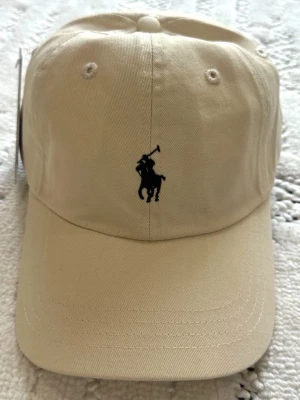 Beige keps från Polo Ralph Lauren - Snygg beige keps från Polo Ralph Lauren med klassisk svart broderad logga framtill och 'POLO' broderat baktill. Justerbar rem bak för perfekt passform. Tillverkad i bomullstyg som ger en avslappnad och stilren look.