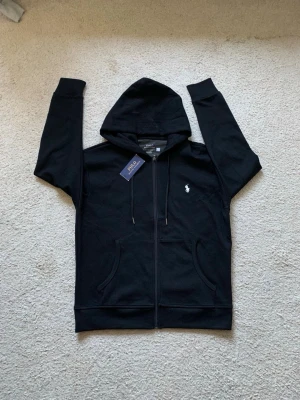 Polo Ralph Lauren hoodie  - Snygg svart hoodie från Polo Ralph Lauren med dragkedja framtill och klassisk logga broderad på bröstet. Hoodien har huva med dragsko och två fickor framtill. Tillverkad i mjukt material som känns skönt mot huden.