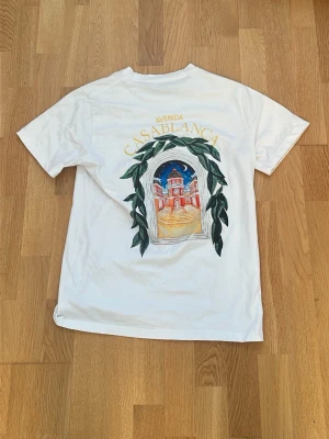 Vit t-shirt med tryck från Casablanca - Vit t-shirt från Casablanca med färgglatt tryck av en byggnad och texten 'Avenida Casablanca' på både fram- och baksidan. T-shirten har korta ärmar och rund hals. Perfekt för dig som gillar unika prints och stilrena plagg.