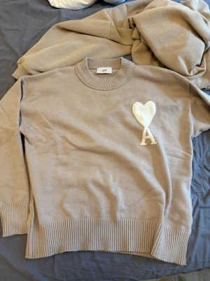 Beige stickad tröja från Ami Paris - Säljer en beige stickad tröja från Ami Paris med rund halsringning och ribbade muddar. På bröstet finns ett vitt hjärta och bokstaven 'A' i samma färg. Perfekt för dig som gillar stilrena och trendiga plagg.